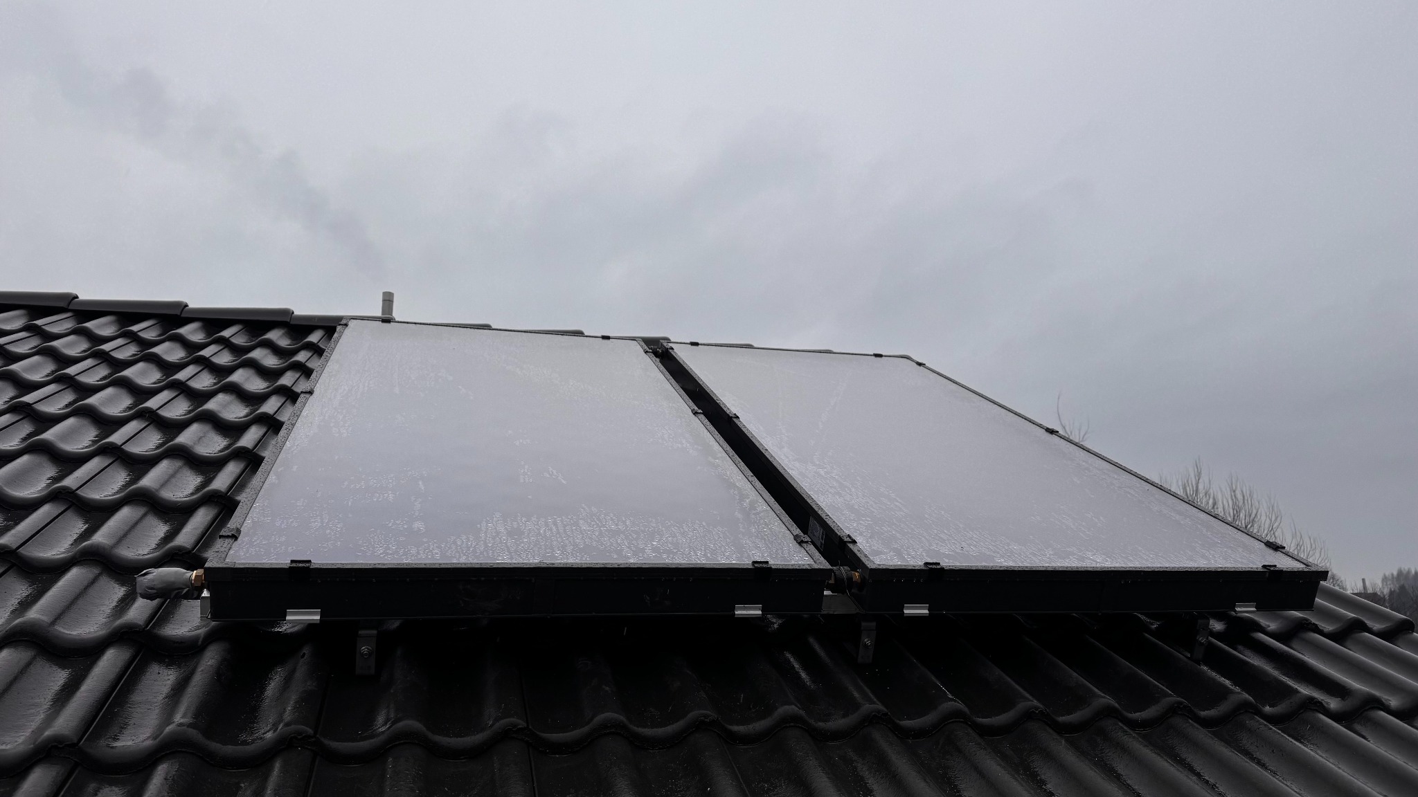 Dwa panele słoneczne, pokryte kroplami wody, zamontowane na czarnym, błyszczącym dachu z dachówki ceramicznej, widoczne elementy montażowe i rura, pochmurne niebo w tle.