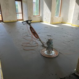 Wlewki maszynowe Bartłomiej Antolak - Szlifowanie wylewki w jasnym, surowym wnętrzu. Widoczne okna z zielenią na zewnątrz, żółta taśma zabezpieczająca przy ścianach. Na podłodze maszyna do wygładzania powierzchni.