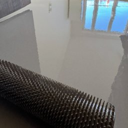 Wlewki maszynowe Bartłomiej Antolak - Wałek kolczasty na świeżo wylanej, szarej posadzce z widocznym odbiciem okna.