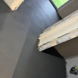 Wlewki maszynowe Bartłomiej Antolak - Szara, gładka powierzchnia podłogi w pomieszczeniu po wylaniu, widok z góry, przy oknie widoczne zielone otoczenie.