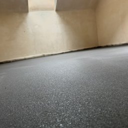 Wlewki maszynowe Bartłomiej Antolak - Świeżo wylana, ciemnoszara posadzka z widoczną fakturą, w pomieszczeniu z surowymi ścianami i oknem dachowym, ujęcie z niskiej perspektywy podkreślające gładkość powierzchni.