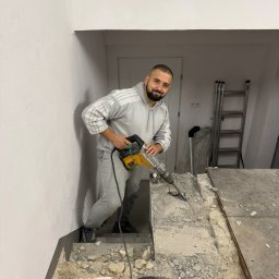 Wlewki maszynowe Bartłomiej Antolak - Uśmiechnięty mężczyzna w szarej bluzie z kapturem i dresach, używa młota udarowego do skuwania starej posadzki w pomieszczeniu w trakcie remontu. W tle widoczne schody i drabina.