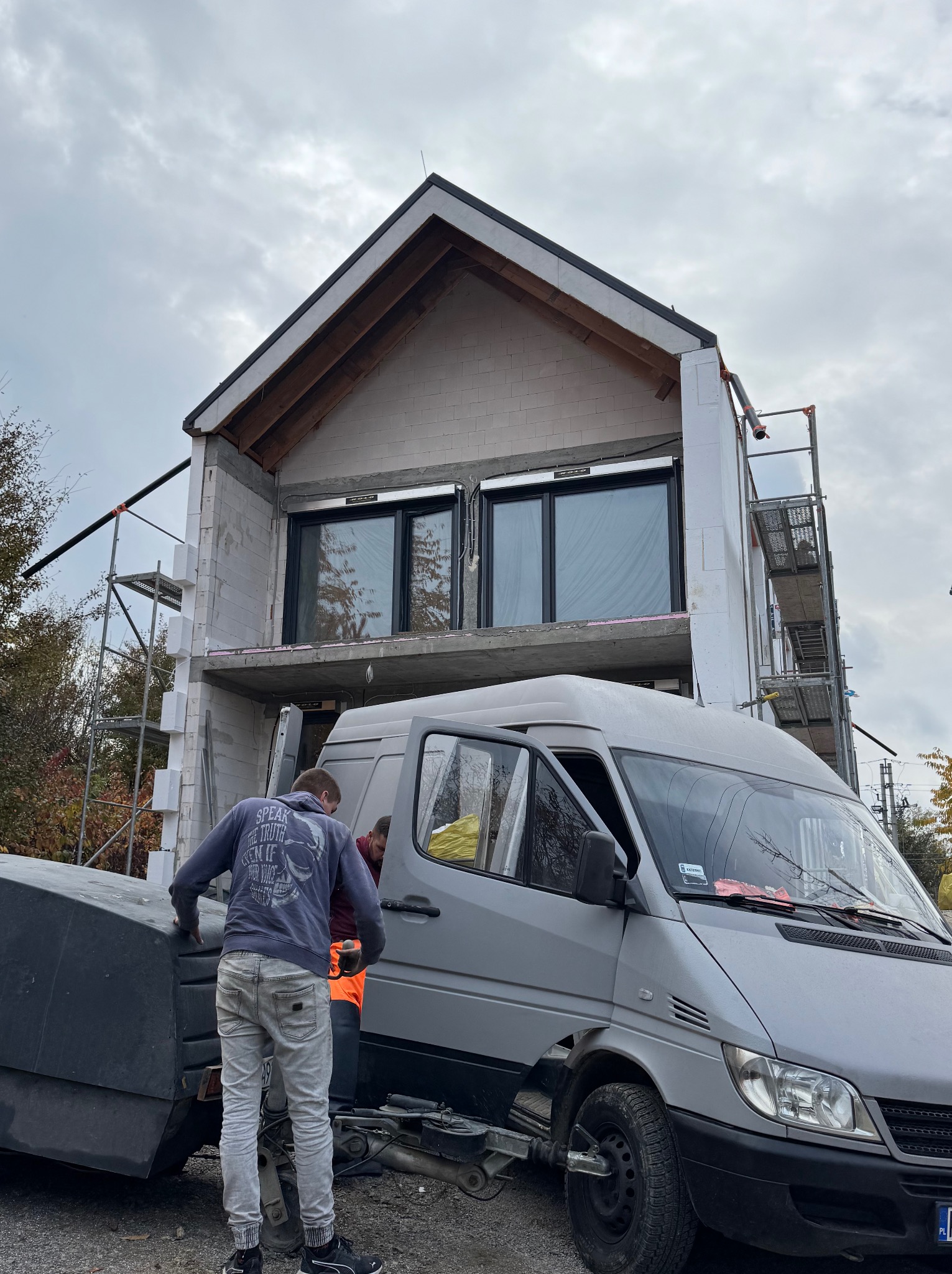 Budowa domu w stanie surowym otwartym z widocznym fragmentem elewacji, ociepleniem styropianowym wokół okien i rusztowaniami, na pierwszym planie pracownicy przy samochodzie dostawczym.