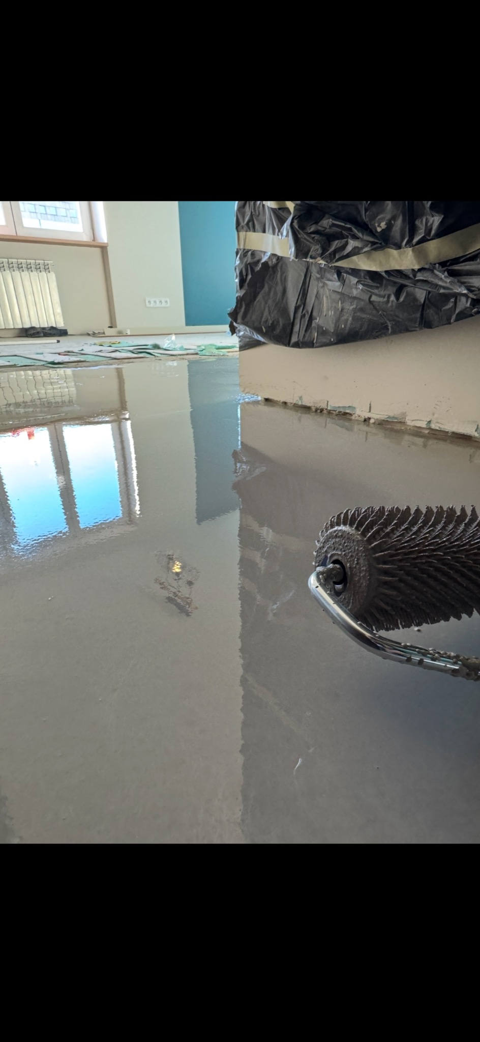 Aplikacja samopoziomującej wylewki na podłodze w pomieszczeniu, widoczny wałek kolczasty do odpowietrzania, ściany zabezpieczone folią malarską.