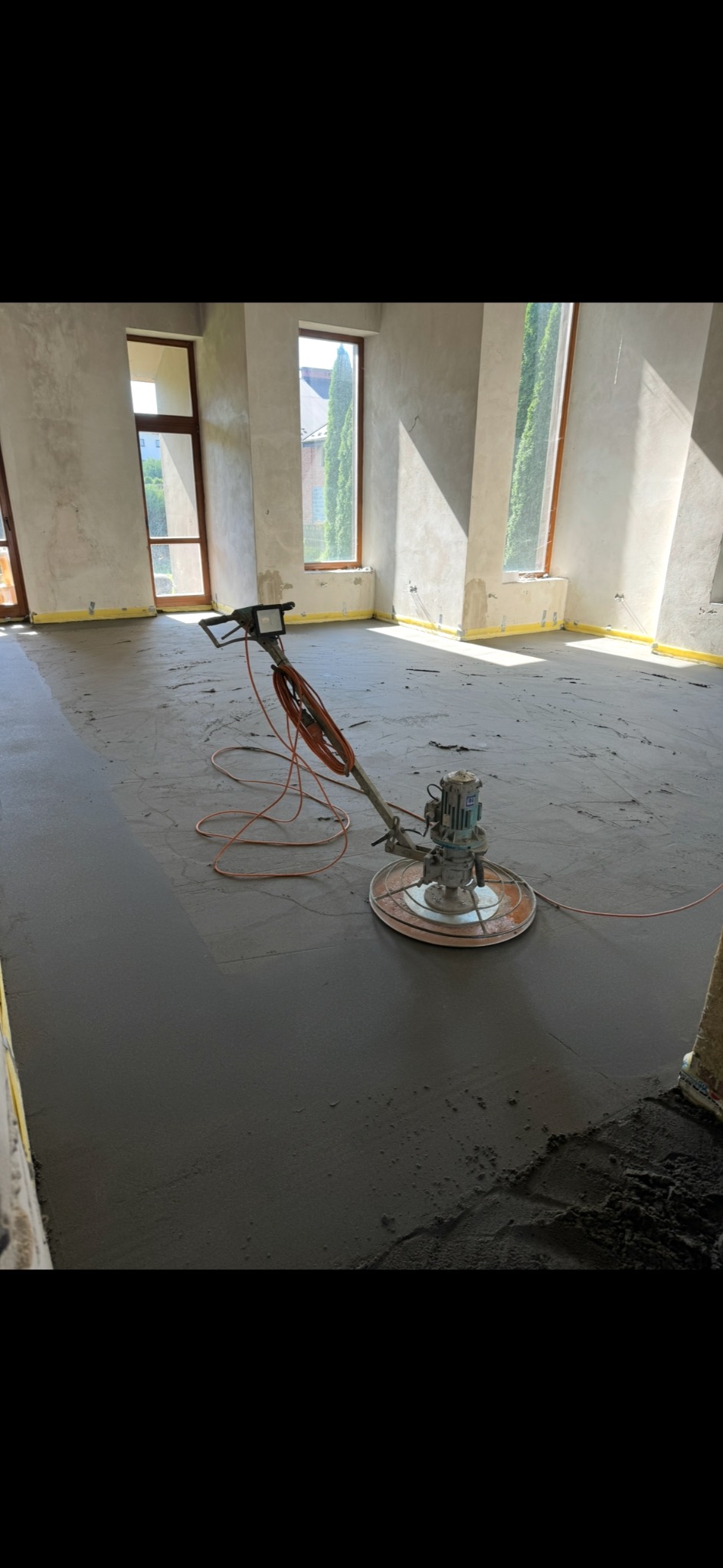 Szlifowanie wylewki w jasnym, surowym wnętrzu. Widoczne okna z zielenią na zewnątrz, żółta taśma zabezpieczająca przy ścianach. Na podłodze maszyna do wygładzania powierzchni.