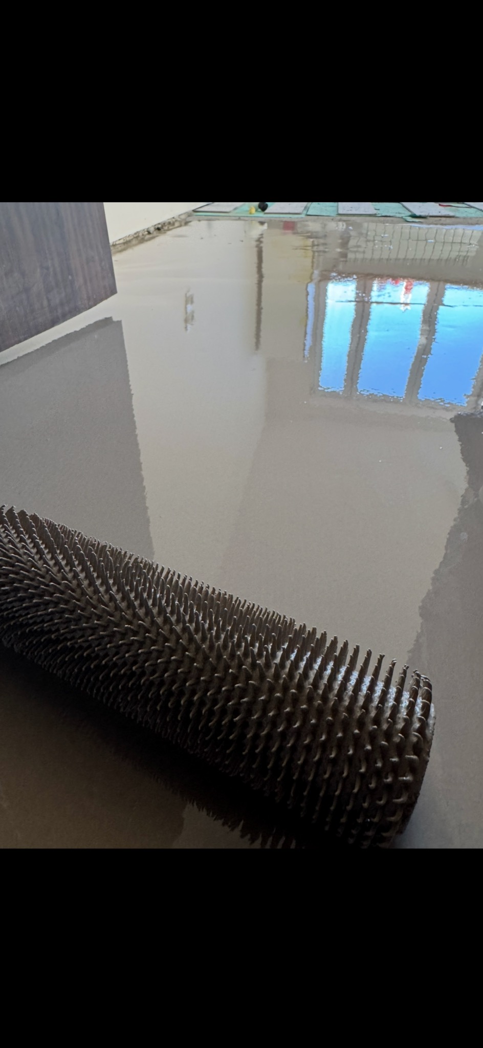 Wałek kolczasty na świeżo wylanej, szarej posadzce z widocznym odbiciem okna.