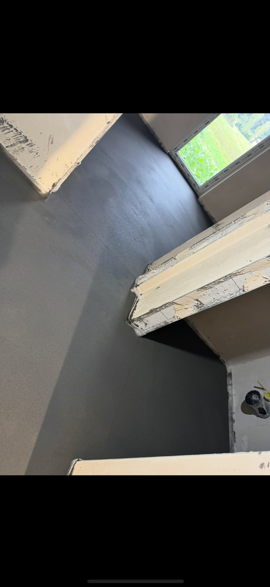 Szara, gładka powierzchnia podłogi w pomieszczeniu po wylaniu, widok z góry, przy oknie widoczne zielone otoczenie.