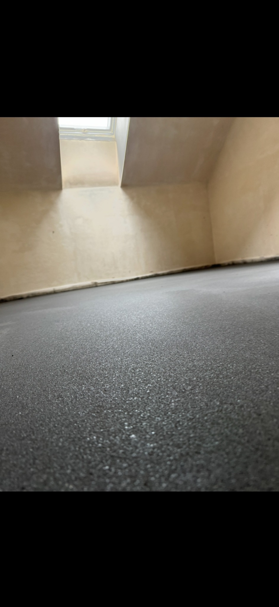 Świeżo wylana, ciemnoszara posadzka z widoczną fakturą, w pomieszczeniu z surowymi ścianami i oknem dachowym, ujęcie z niskiej perspektywy podkreślające gładkość powierzchni.