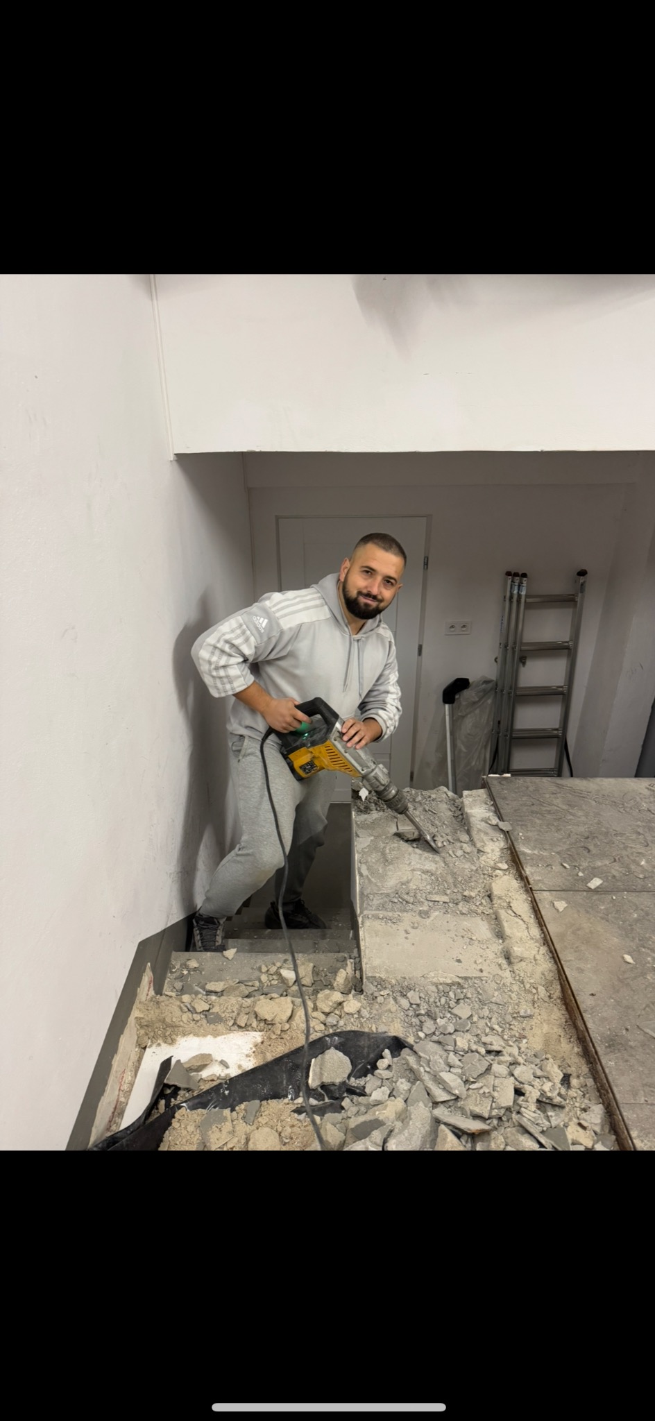 Uśmiechnięty mężczyzna w szarej bluzie z kapturem i dresach, używa młota udarowego do skuwania starej posadzki w pomieszczeniu w trakcie remontu. W tle widoczne schody i drabina.