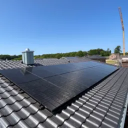 Czarny panel słoneczny na ciemnobrązowym dachu z dachówki ceramicznej, w tle błękitne niebo i żuraw budowlany.