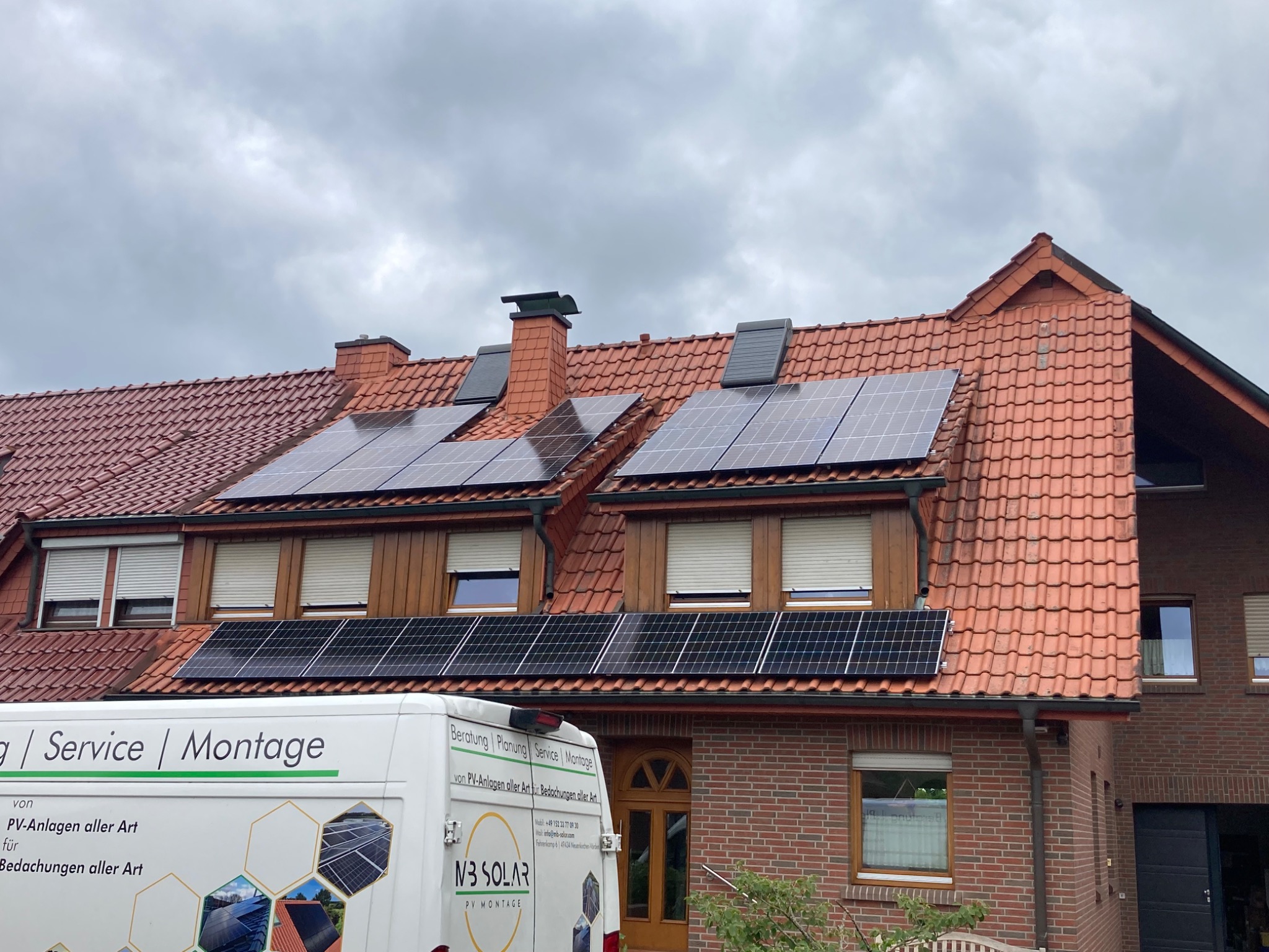 Czerwony dach z zamontowanymi panelami słonecznymi, widoczny fragment firmowego samochodu z logiem i hasłem 'PV Montage'.