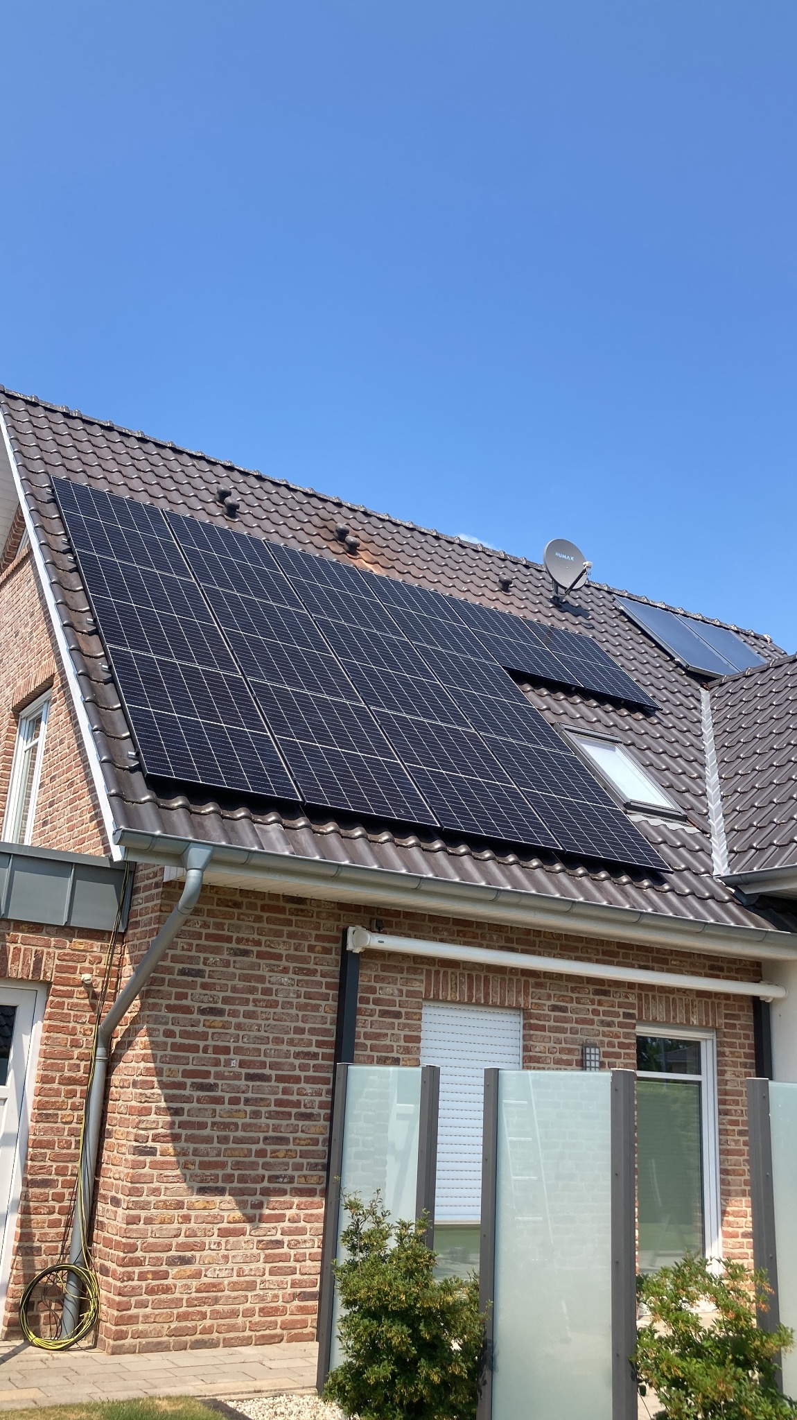 Panele fotowoltaiczne na brązowym dachu domu z cegły, widoczne okno dachowe i antena satelitarna, błękitne niebo w tle.