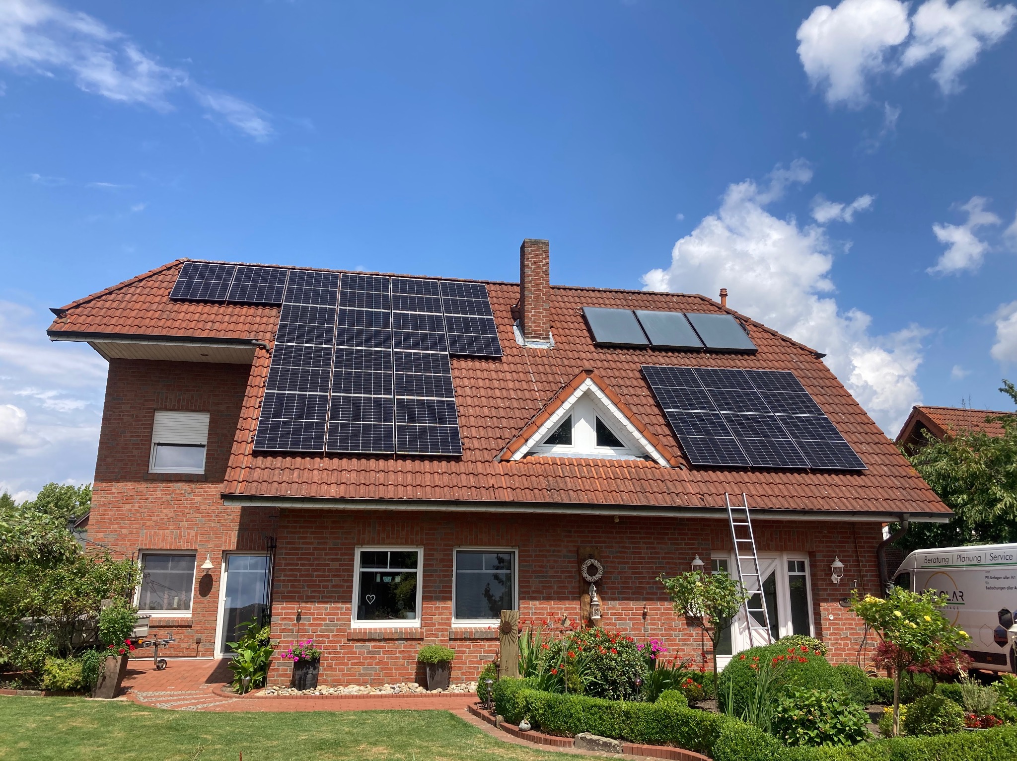 Czerwony dom z cegły z panelami słonecznymi na dachu i srebrną drabiną opartą o ścianę. Zadbane rabaty kwiatowe i trawnik przed budynkiem. Na podjeździe zaparkowany van firmy instalującej panele.