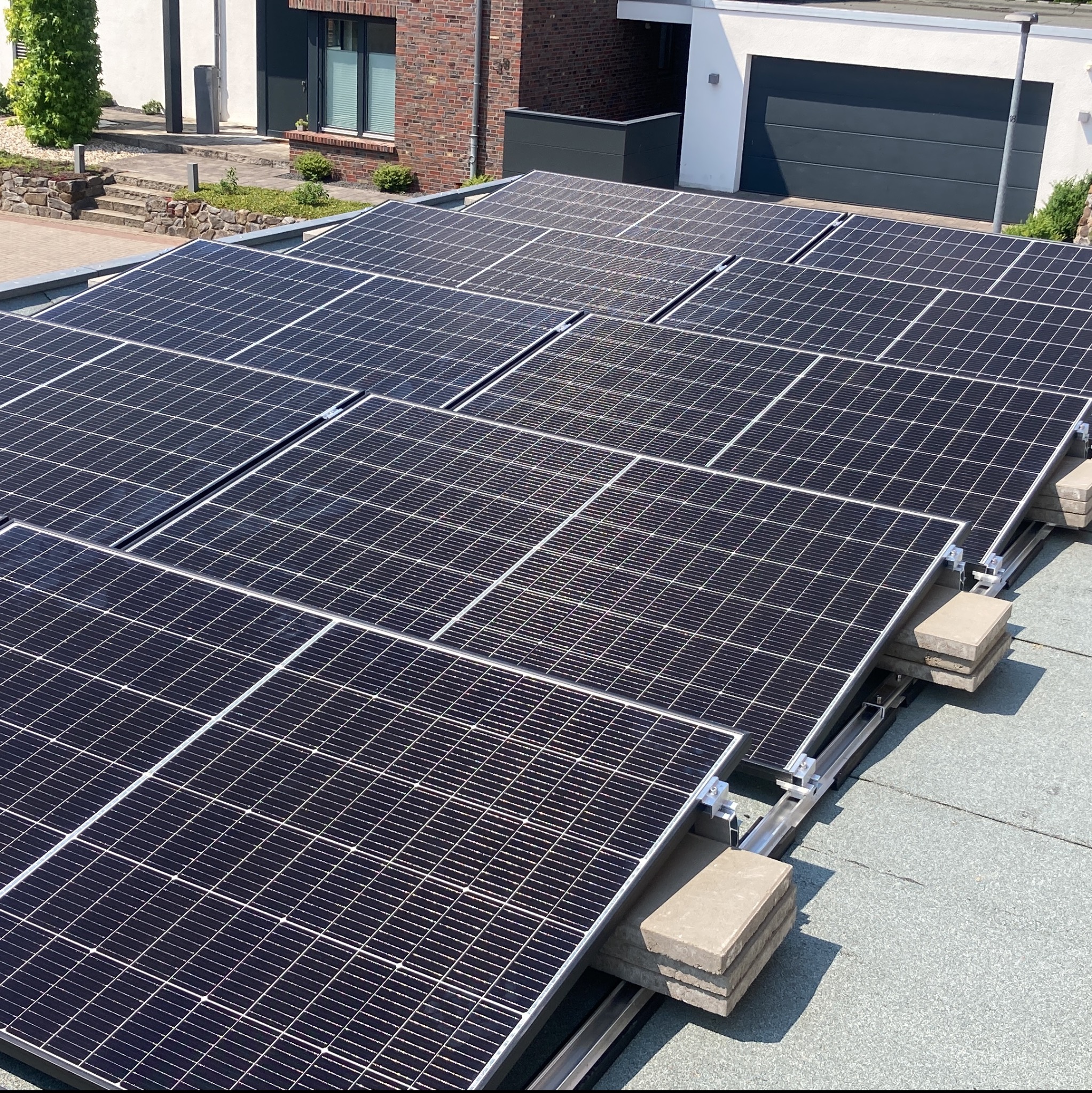 Panele słoneczne zamontowane na płaskim dachu budynku mieszkalnego, widoczne betonowe obciążniki i metalowe elementy montażowe.