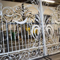 Biała, dekoracyjna brama z kutego żelaza z motywami roślinnymi, w trakcie produkcji w warsztacie, z widocznymi maszynami w tle.