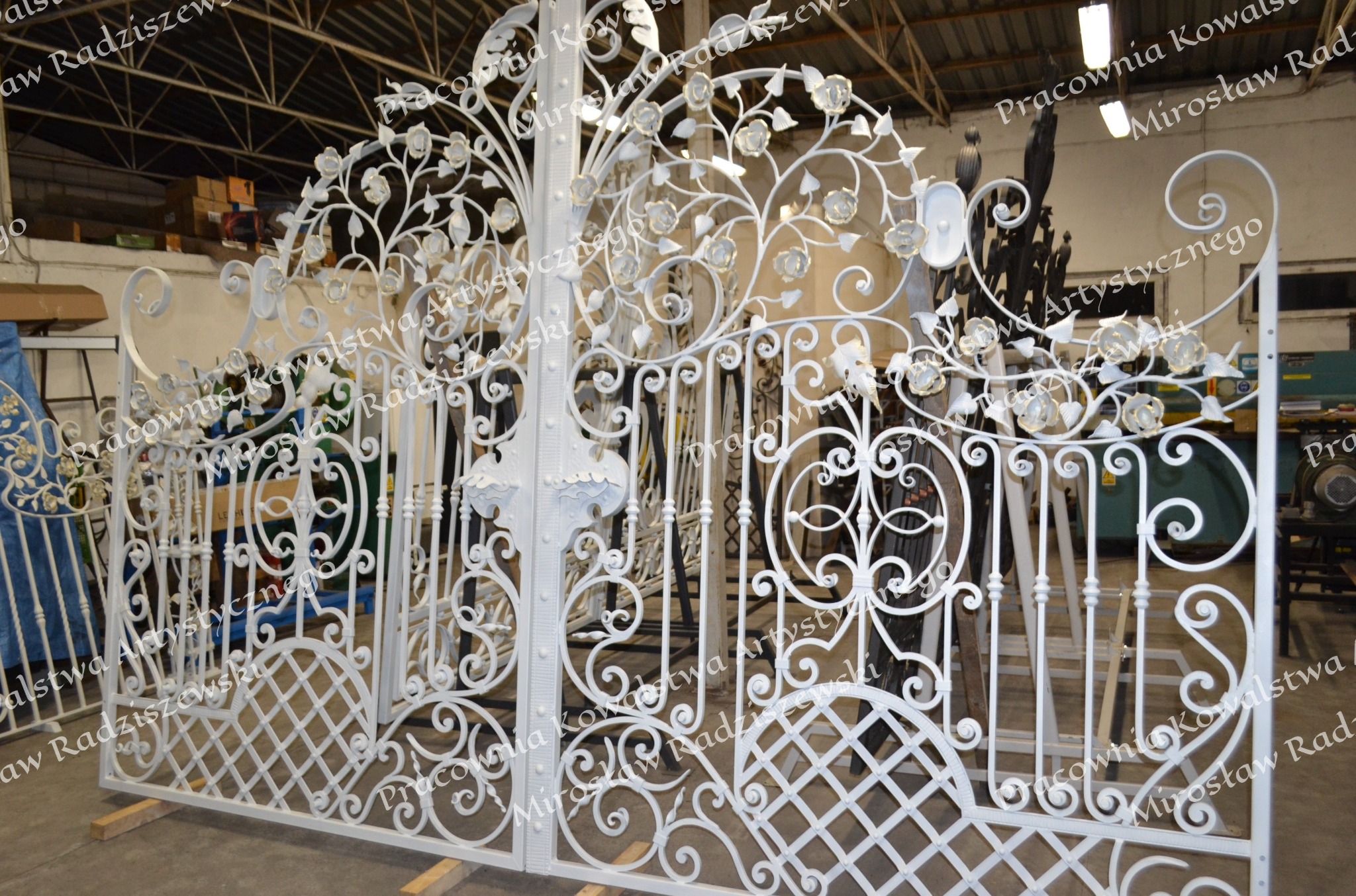 Biała brama kuta z motywem roślinnym, w warsztacie, widoczne elementy dekoracyjne w kształcie róż i liści, z siatkowym wypełnieniem dolnej części.