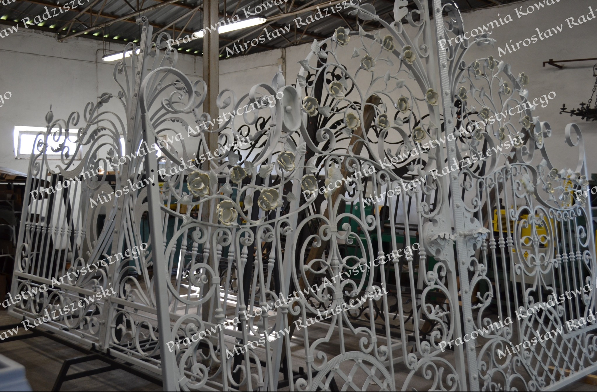 Biała, ręcznie kuta brama wjazdowa z motywem kwiatowym róży, widoczna w warsztacie przed montażem.