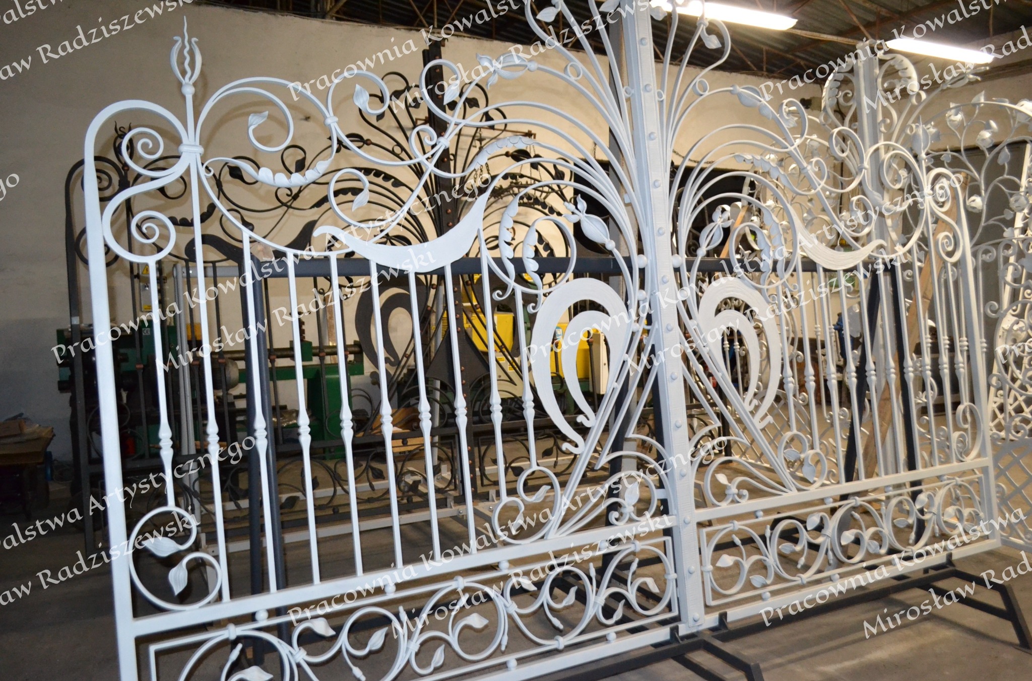 Biała, dekoracyjna brama z kutego żelaza z motywami roślinnymi, w trakcie produkcji w warsztacie, z widocznymi maszynami w tle.
