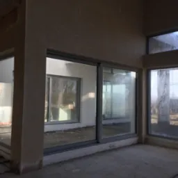 Surowe wnętrze w trakcie prac wykończeniowych, widoczne ślady po nałożeniu tynku na ścianach, duże okna z widokiem na zewnątrz, podłoga betonowa, brak wykończenia.