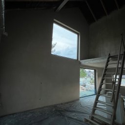 Dmytro Lonchak - Wnętrze w trakcie prac wykończeniowych: ściany pokryte tynkiem, okna z widokiem na zewnątrz, drabina oparta o ścianę. Podłoga zabezpieczona folią.