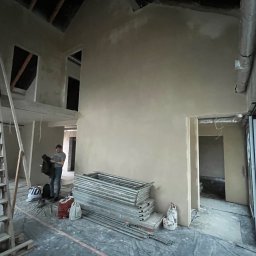 Dmytro Lonchak - Wnętrze w trakcie prac wykończeniowych: świeżo otynkowane ściany, rozłożone rusztowania, widoczna osoba w tle. Surowy stan, przygotowanie do dalszych etapów.
