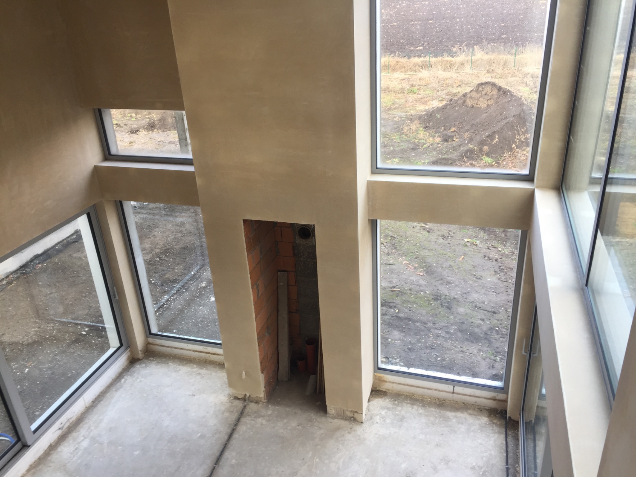 Surowe wnętrze w stanie deweloperskim z dużymi oknami i ścianami pokrytymi świeżym tynkiem w odcieniach beżu, widoczny fragment ściany z cegły w miejscu na kominek.