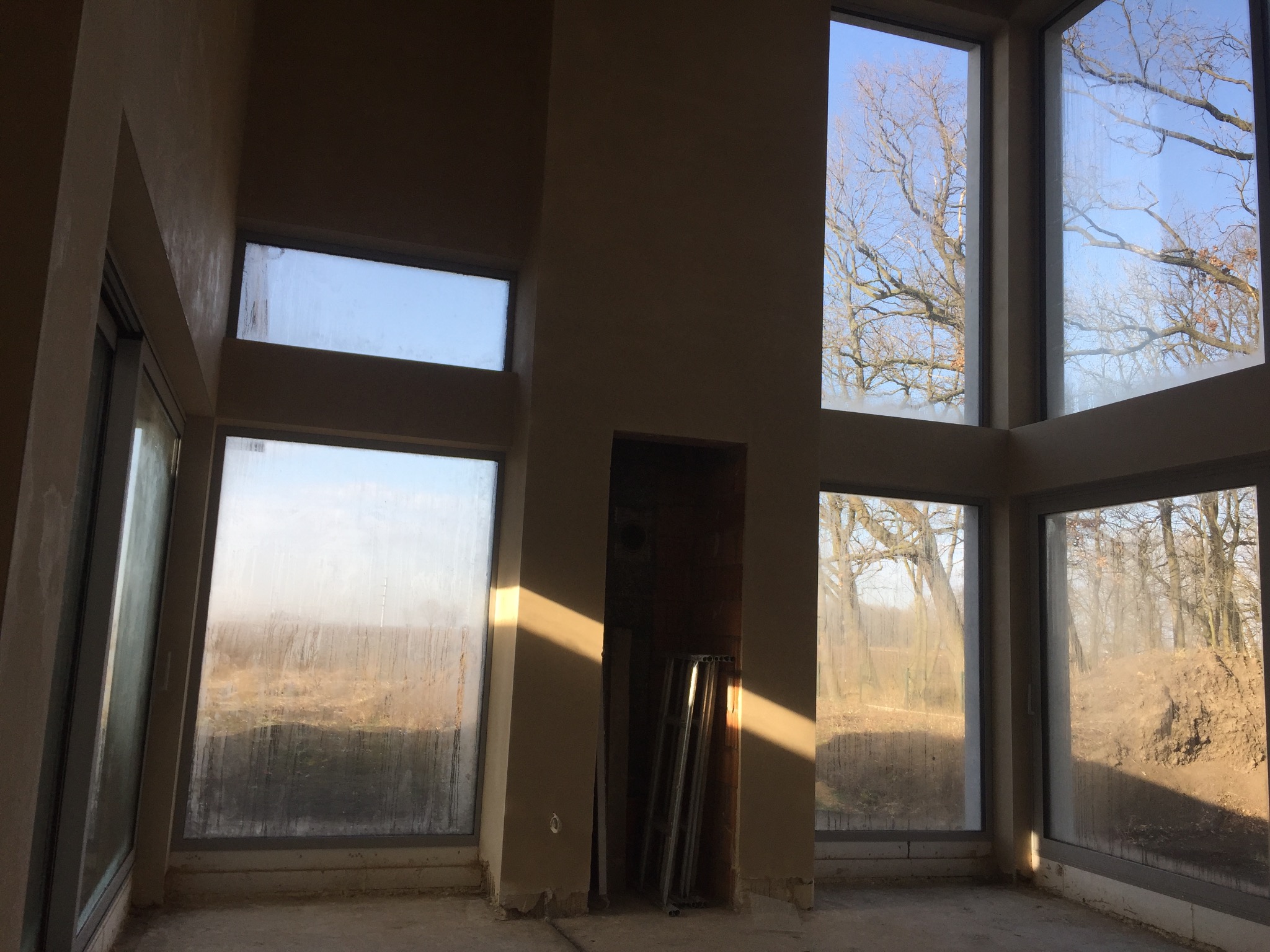 Surowy stan wnętrza z dużymi oknami, widoczne ślady wilgoci na szybach, fragment ściany z nałożoną warstwą tynku, wnęka z opartymi metalowymi profilami.