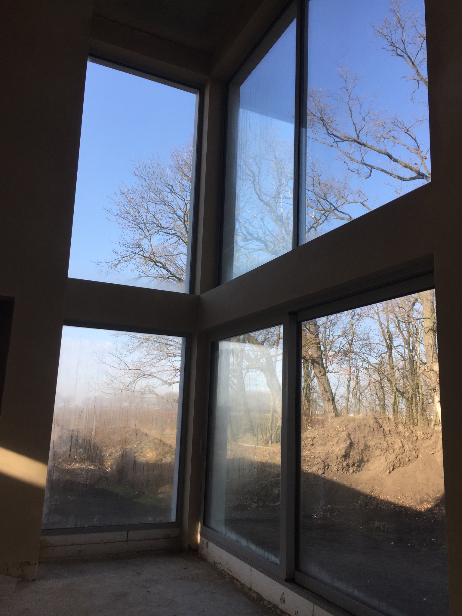 Wnętrze w stanie surowym z dużymi oknami, przez które widać drzewa i ziemię, ściany przygotowane do prac wykończeniowych.