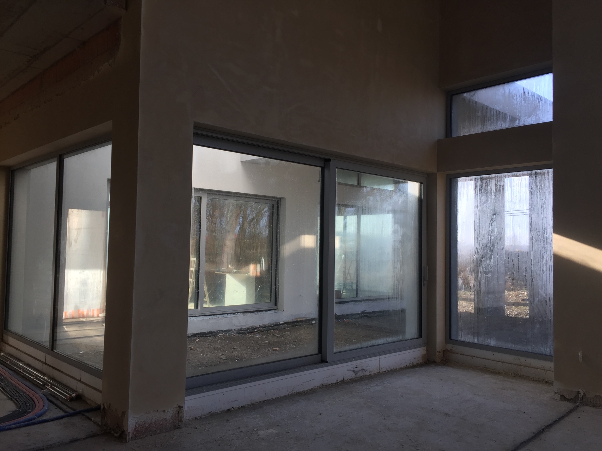 Surowe wnętrze w trakcie prac wykończeniowych, widoczne ślady po nałożeniu tynku na ścianach, duże okna z widokiem na zewnątrz, podłoga betonowa, brak wykończenia.