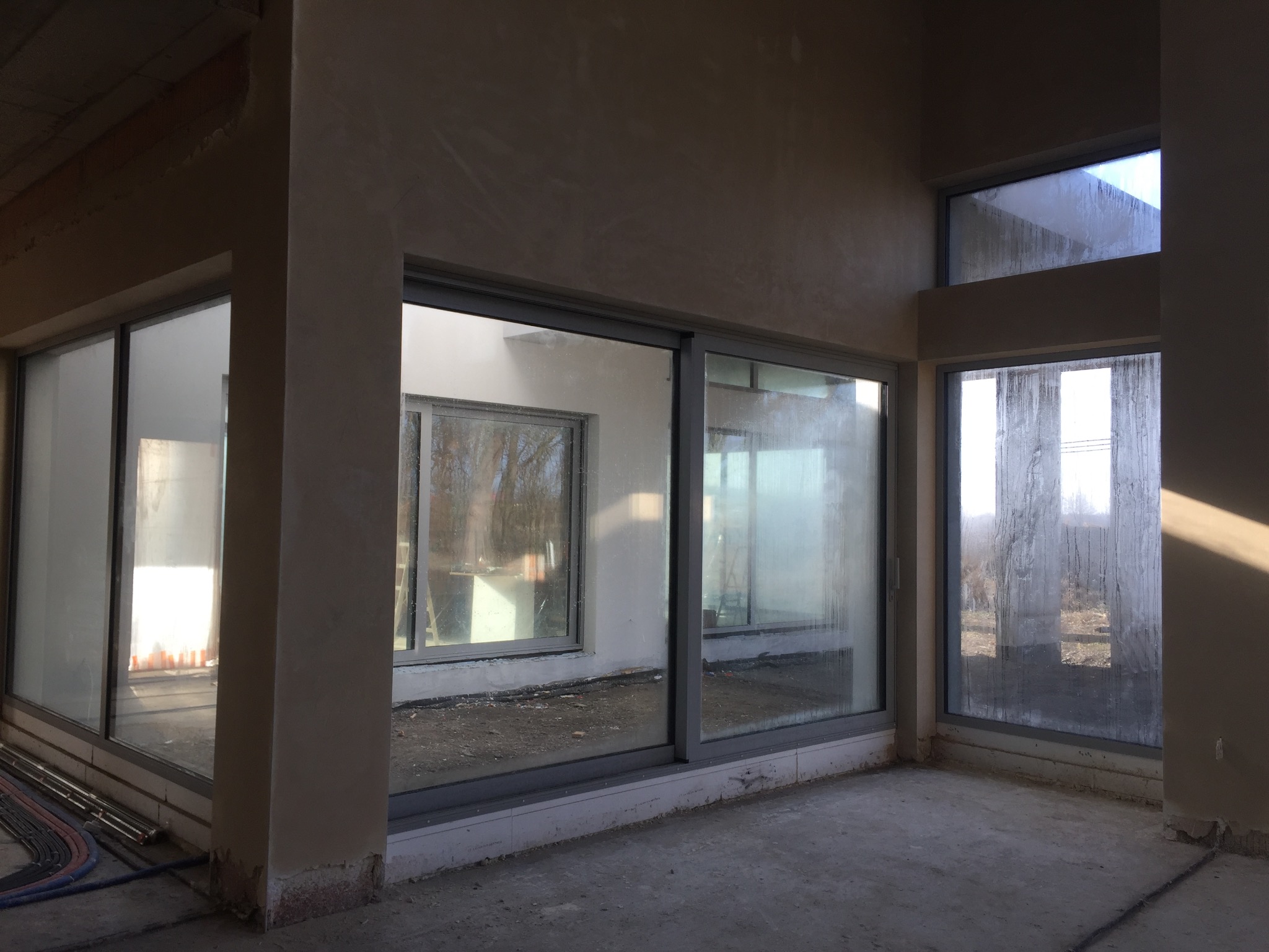 Surowe wnętrze w trakcie prac wykończeniowych; ściany pokryte świeżym tynkiem, duże okna z widokiem na zewnątrz, widoczne ślady wilgoci na szybach i niedoskonałości na powierzchni tynku.