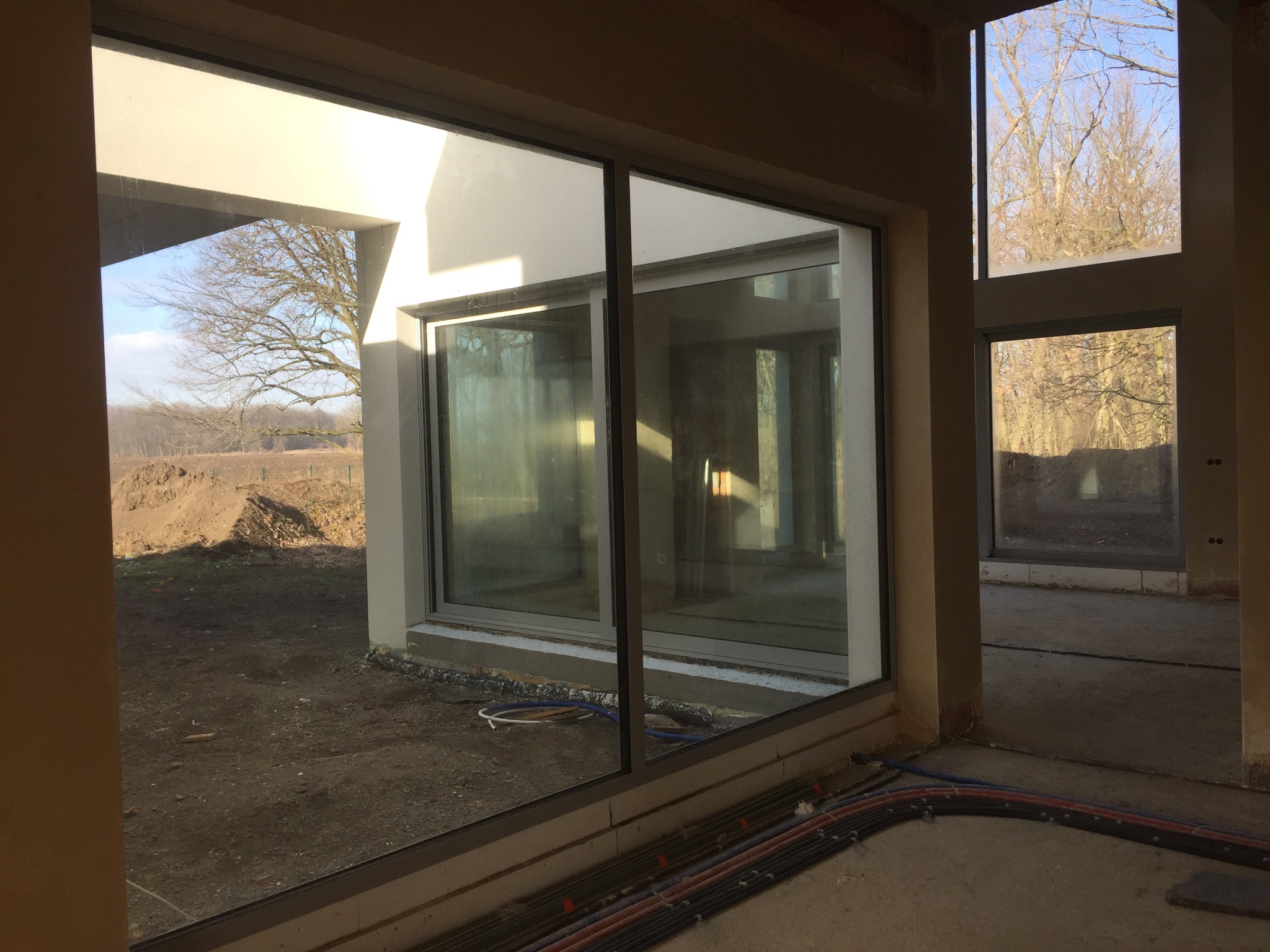 Wnętrze w stanie surowym z dużymi oknami aluminiowymi, widok na zewnątrz na ziemię i drzewa bez liści, widoczne kable instalacyjne na podłodze wewnątrz budynku.