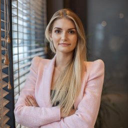 Beflawless Clinic EDYTA WOJEWÓDZKA - Portret kobiety z blond włosami w różowym żakiecie, stojącej przy oknie z żaluzjami, skrzyżowane ręce, delikatny makijaż.