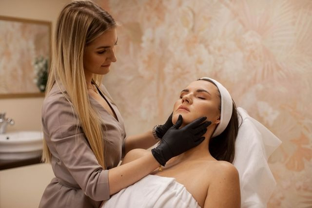 Kobieta z zamkniętymi oczami leży na leżance podczas zabiegu pielęgnacyjnego twarzy wykonywanego przez kosmetyczkę w czarnych rękawiczkach, widoczne jasne włosy kosmetyczki i delikatne tło w salonie.