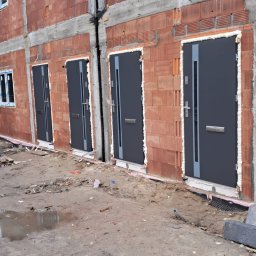 S.V.MAX.PL SPÓŁKA Z OGRANICZONĄ ODPOWIEDZIALNOŚCIĄ - Szereg domów w trakcie budowy z zamontowanymi szarymi drzwiami wejściowymi, widoczne pianowanie wokół ościeżnic, surowy stan elewacji z czerwonej cegły dziurawki.