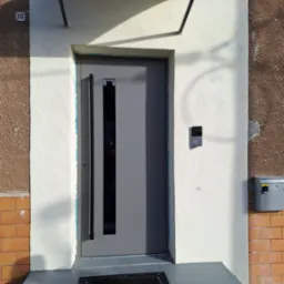 Szare drzwi wejściowe z długim, pionowym uchwytem i małym przeszkleniem, zamontowane w ościeżnicy z kremowego tynku pod daszkiem, z numerem domu '10' po lewej stronie i domofonem po prawej.