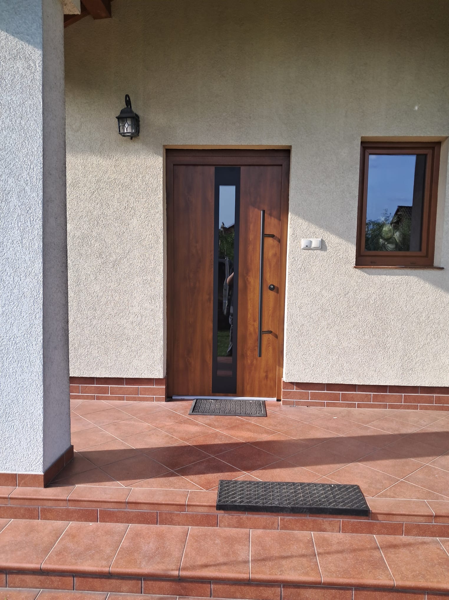 Brązowe drzwi wejściowe z pionowym przeszkleniem i długim uchwytem, zamontowane w jasnym budynku z małym oknem po prawej stronie; przed drzwiami wycieraczka na brązowych płytkach.