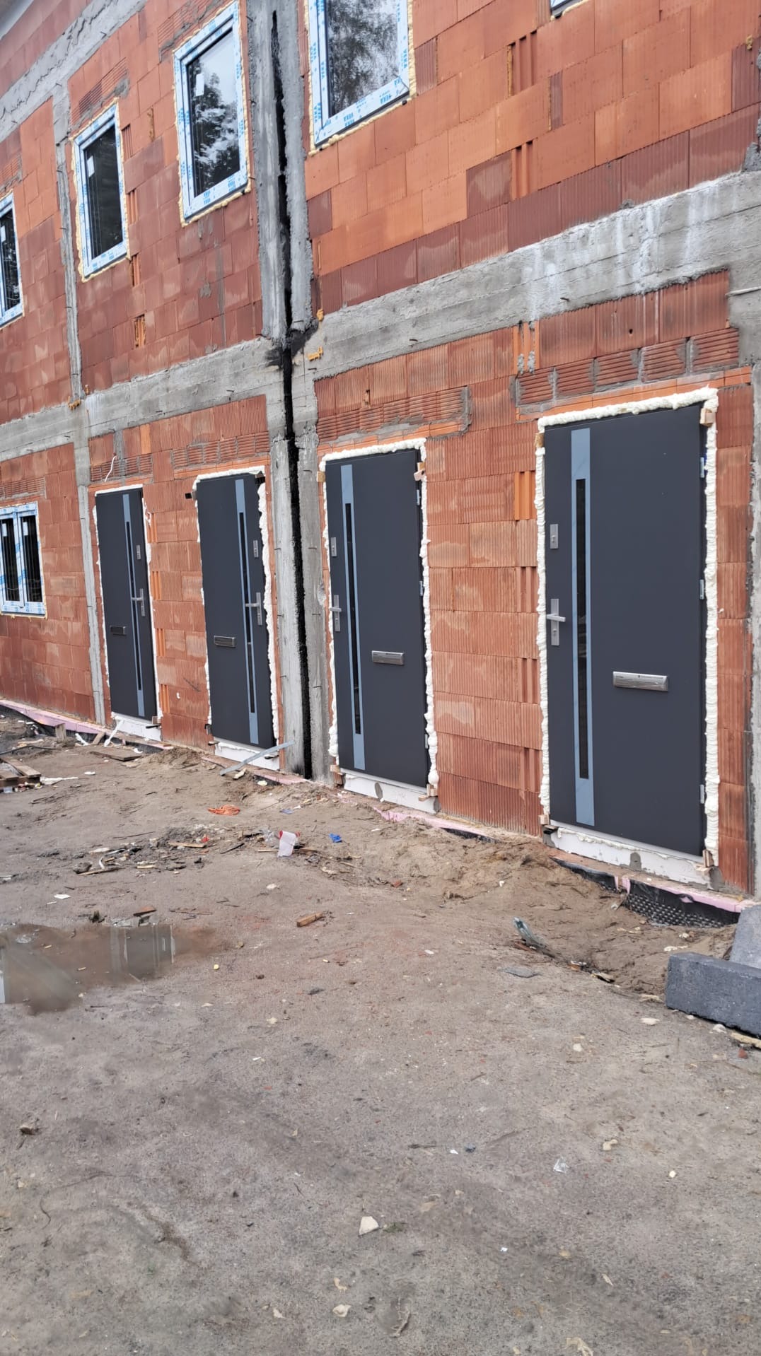 Szereg domów w trakcie budowy z zamontowanymi szarymi drzwiami wejściowymi, widoczne pianowanie wokół ościeżnic, surowy stan elewacji z czerwonej cegły dziurawki.