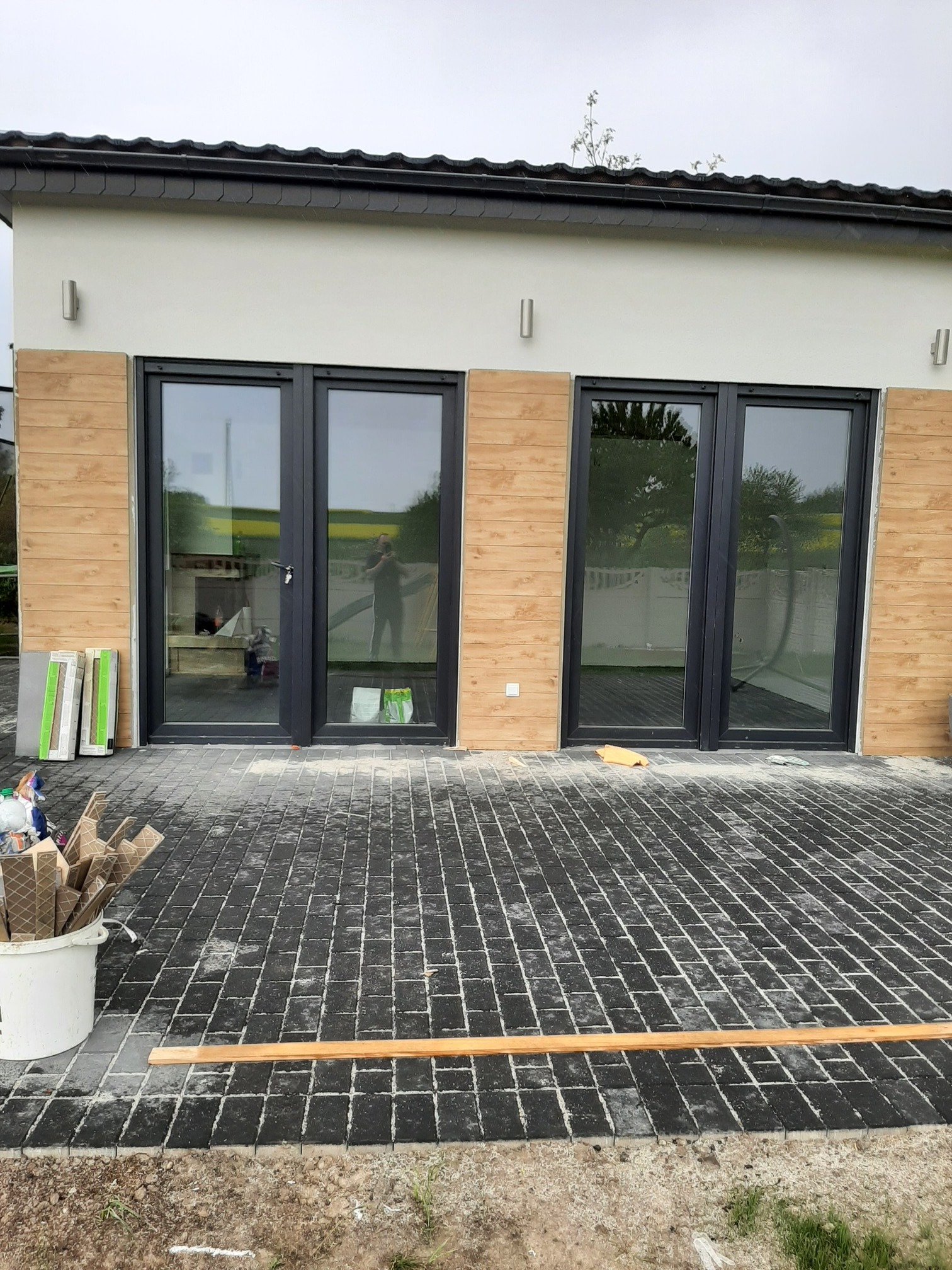 Dwa zestawy dwuskrzydłowych drzwi zewnętrznych z czarnymi ramami, zamontowane w budynku z elewacją w odcieniach beżu i brązu, Opole. Na pierwszym planie kostka brukowa, ślady prac montażowych...