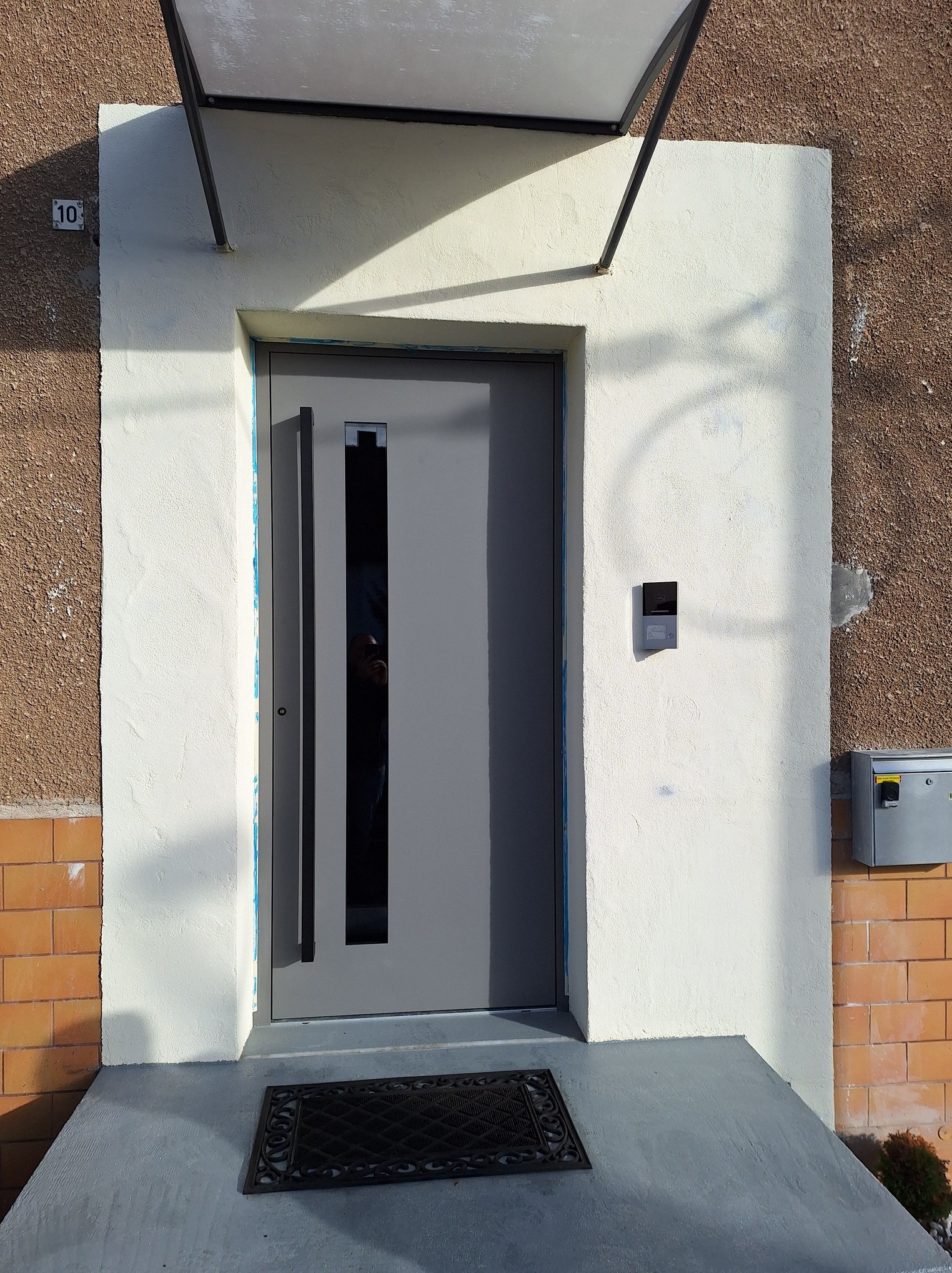 Szare drzwi wejściowe z długim, pionowym uchwytem i małym przeszkleniem, zamontowane w ościeżnicy z kremowego tynku pod daszkiem, z numerem domu '10' po lewej stronie i domofonem po prawej.