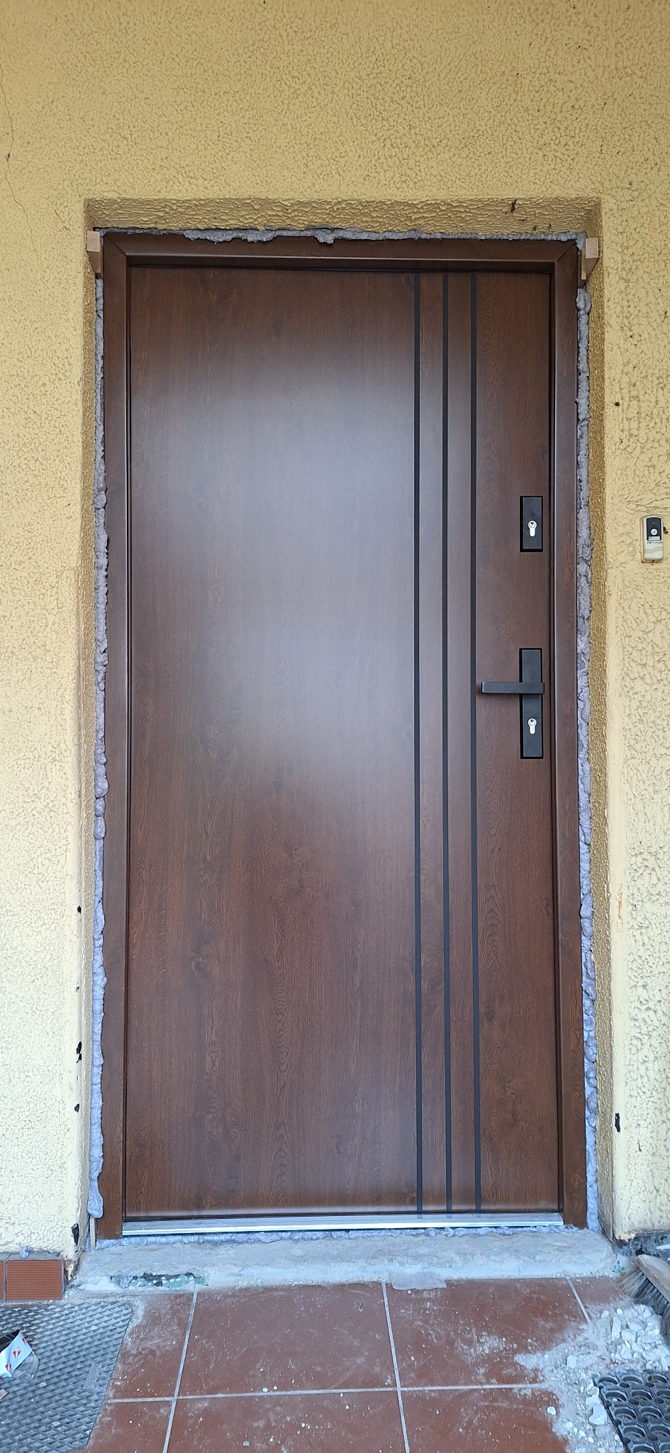 Brązowe drzwi wejściowe z trzema pionowymi, czarnymi paskami i czarną klamką, osadzone w ościeżnicy z widoczną pianką montażową, na tle beżowej ściany o chropowatej fakturze i częściowo wykończonej...