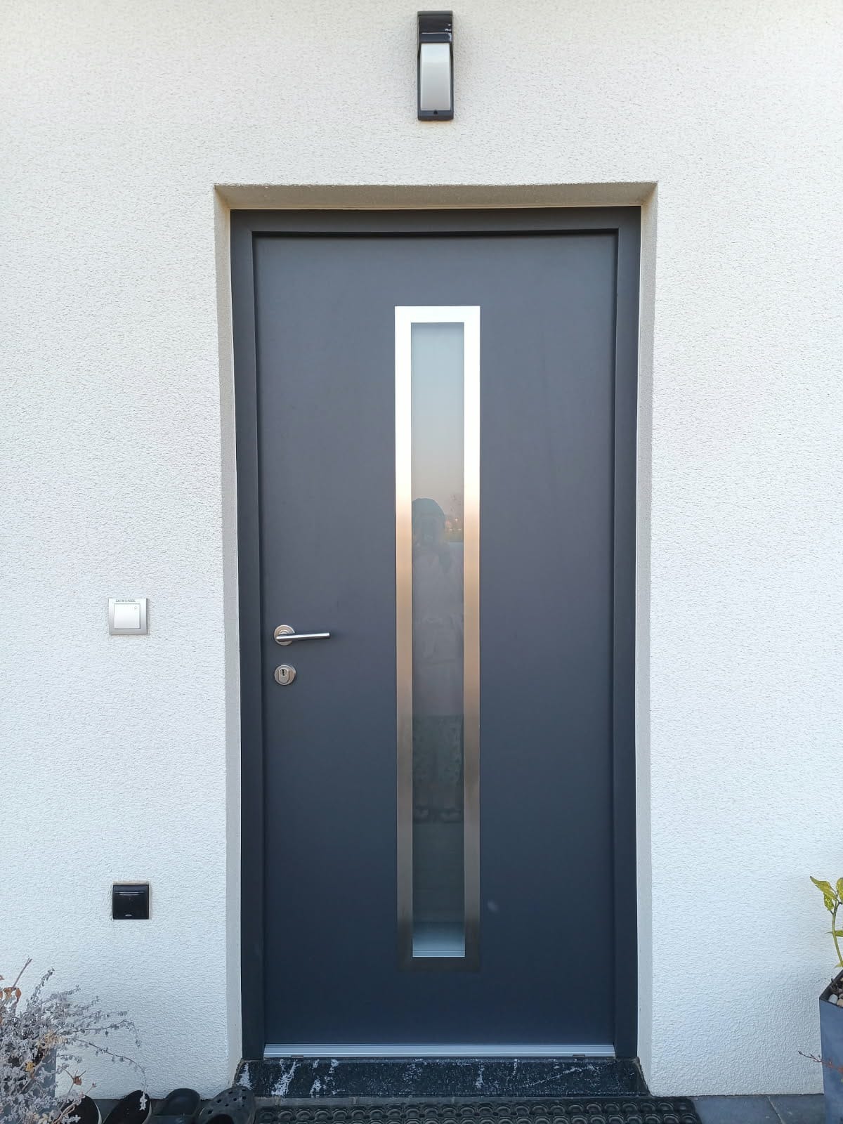 Grafitowe drzwi wejściowe z pionowym przeszkleniem w metalowej ramie, zamontowane w białej, teksturowanej ścianie budynku z widocznym oświetleniem zewnętrznym i wycieraczką.