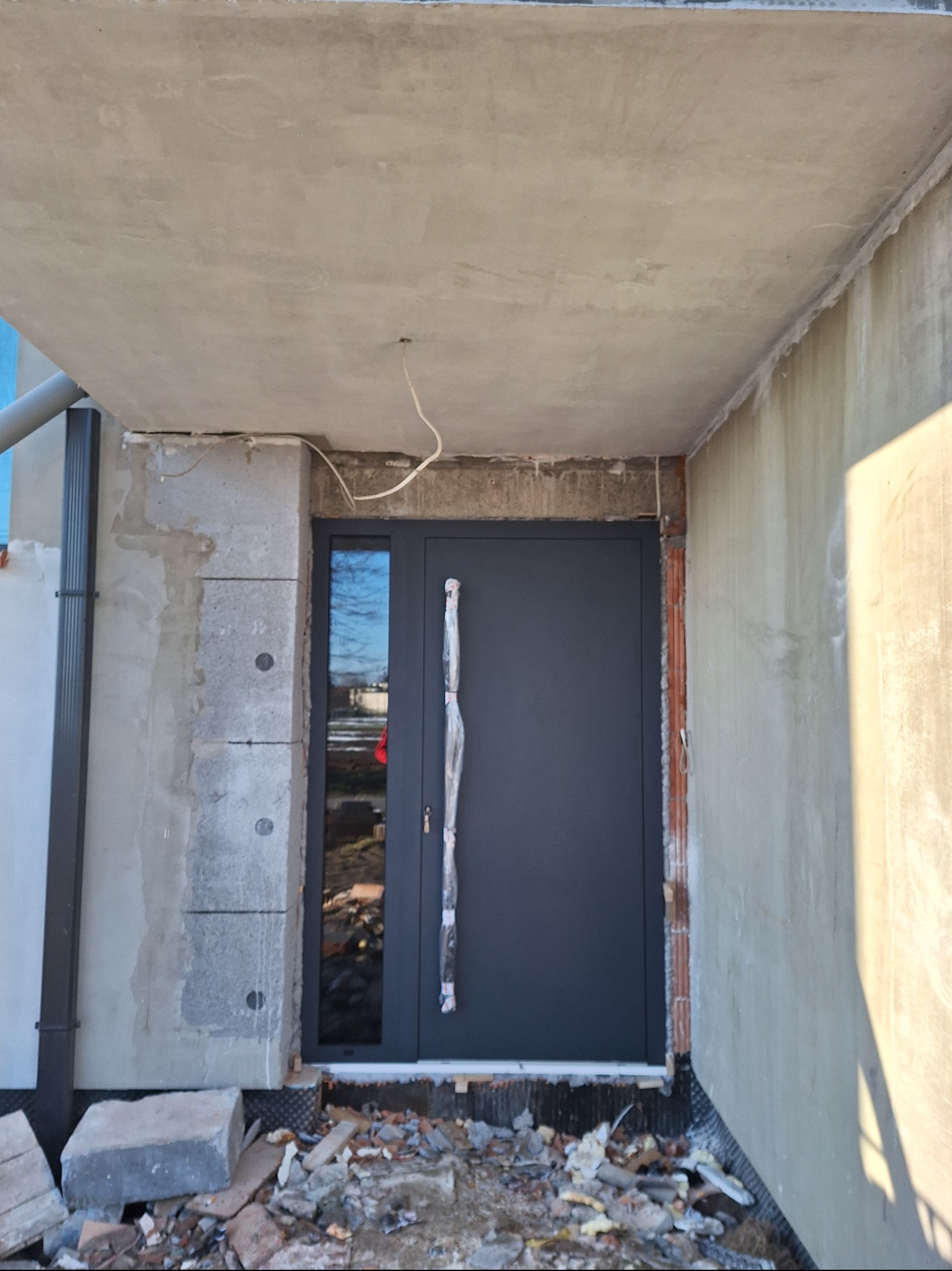 Ciemnoszare drzwi zewnętrzne z szybą boczną, z folią ochronną na uchwycie, zamontowane w surowym stanie budynku, widoczne niedokończone ściany i gruz na podłożu.