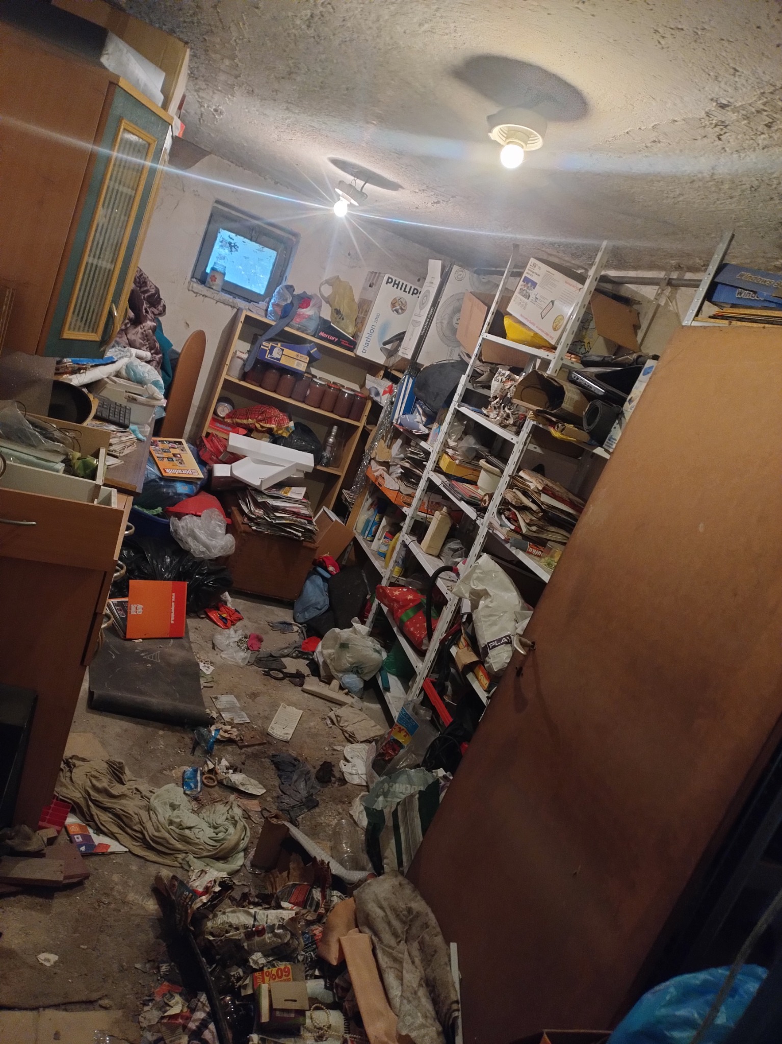 Zagracony pokój z półkami, kartonami, starymi meblami i stertami śmieci na podłodze, oświetlony trzema żarówkami, widok z ukosa.