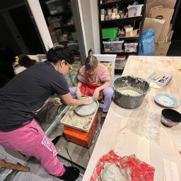 Pracownia Art Terapii - Kobieta z dziewczynką formują glinę na kole garncarskim w warsztacie, widoczny kubeł z gliną, narzędzia i gotowe wyroby.