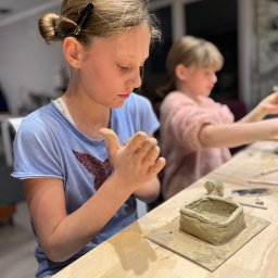 Pracownia Art Terapii - Dziewczynka formuje glinę przy drewnianym stole, tworząc małe naczynie; w tle druga dziewczynka również pracuje z gliną.