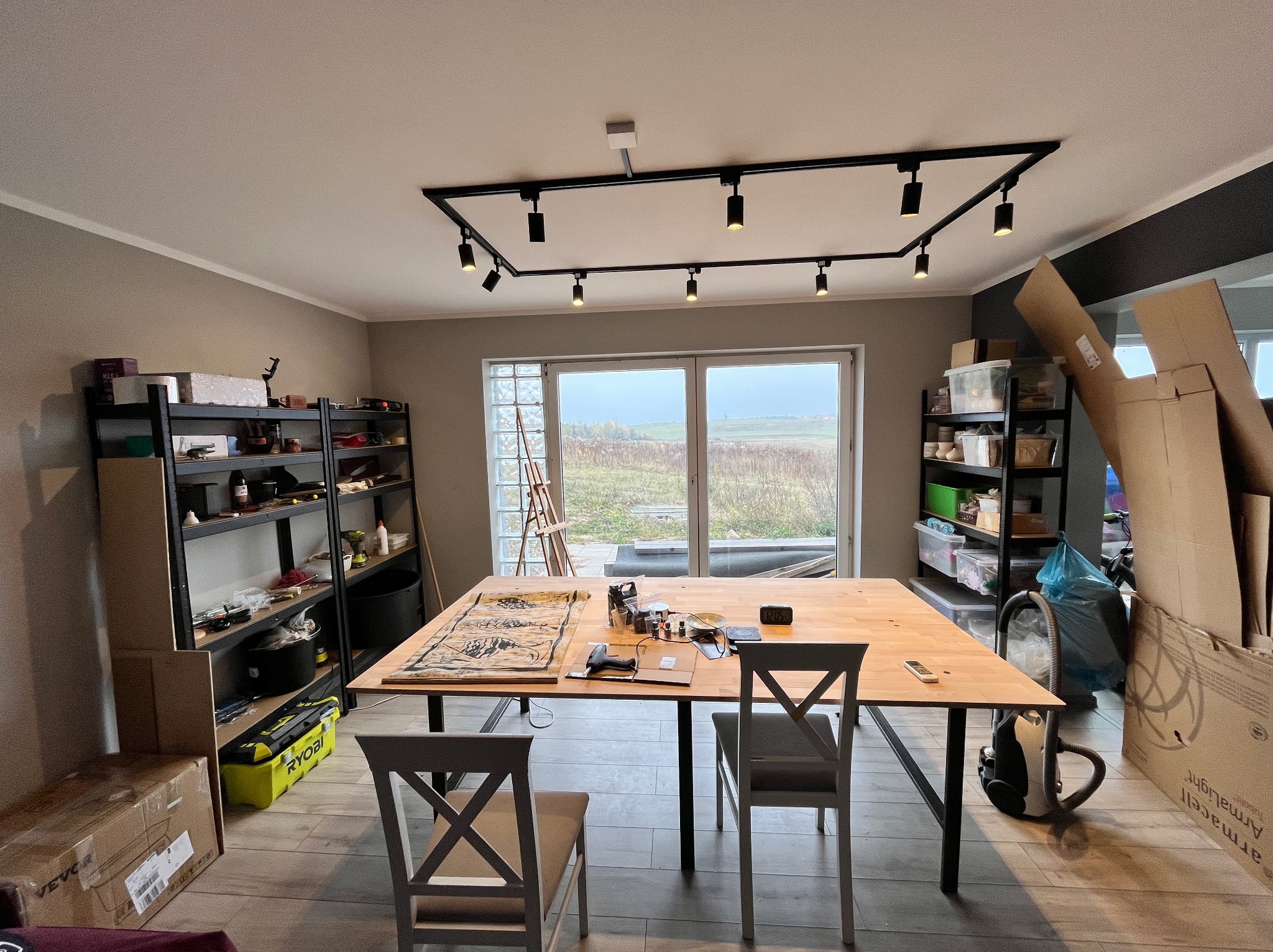 Pracownia z dużym stołem roboczym, na którym leży rozłożony wzór do malowania, narzędzia, oświetlenie sufitowe, półki z akcesoriami i kartonami w tle.