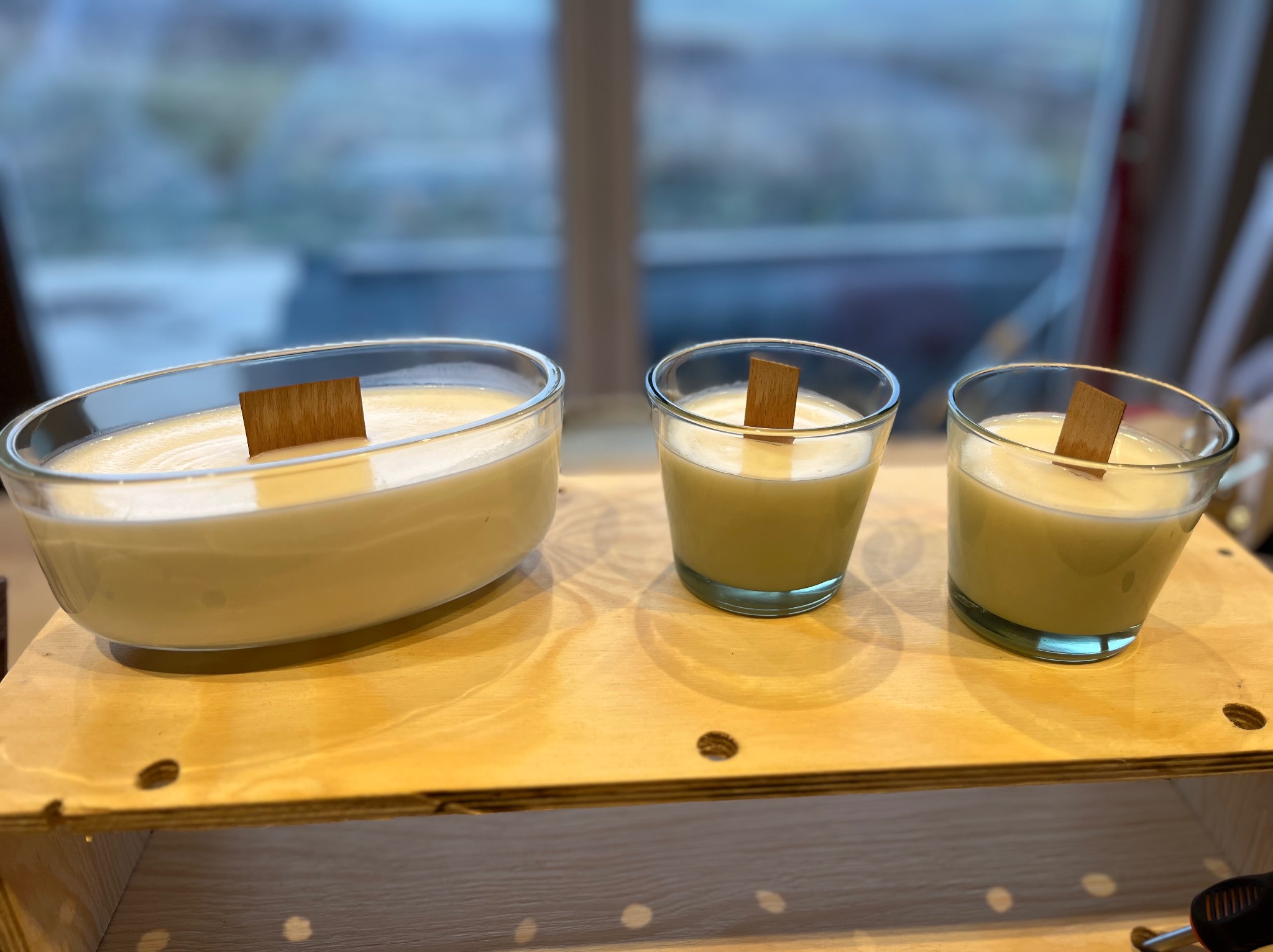 Trzy świece sojowe z drewnianymi knotami w szklanych naczyniach o różnej wielkości, ustawione na surowej drewnianej półce na tle okna.