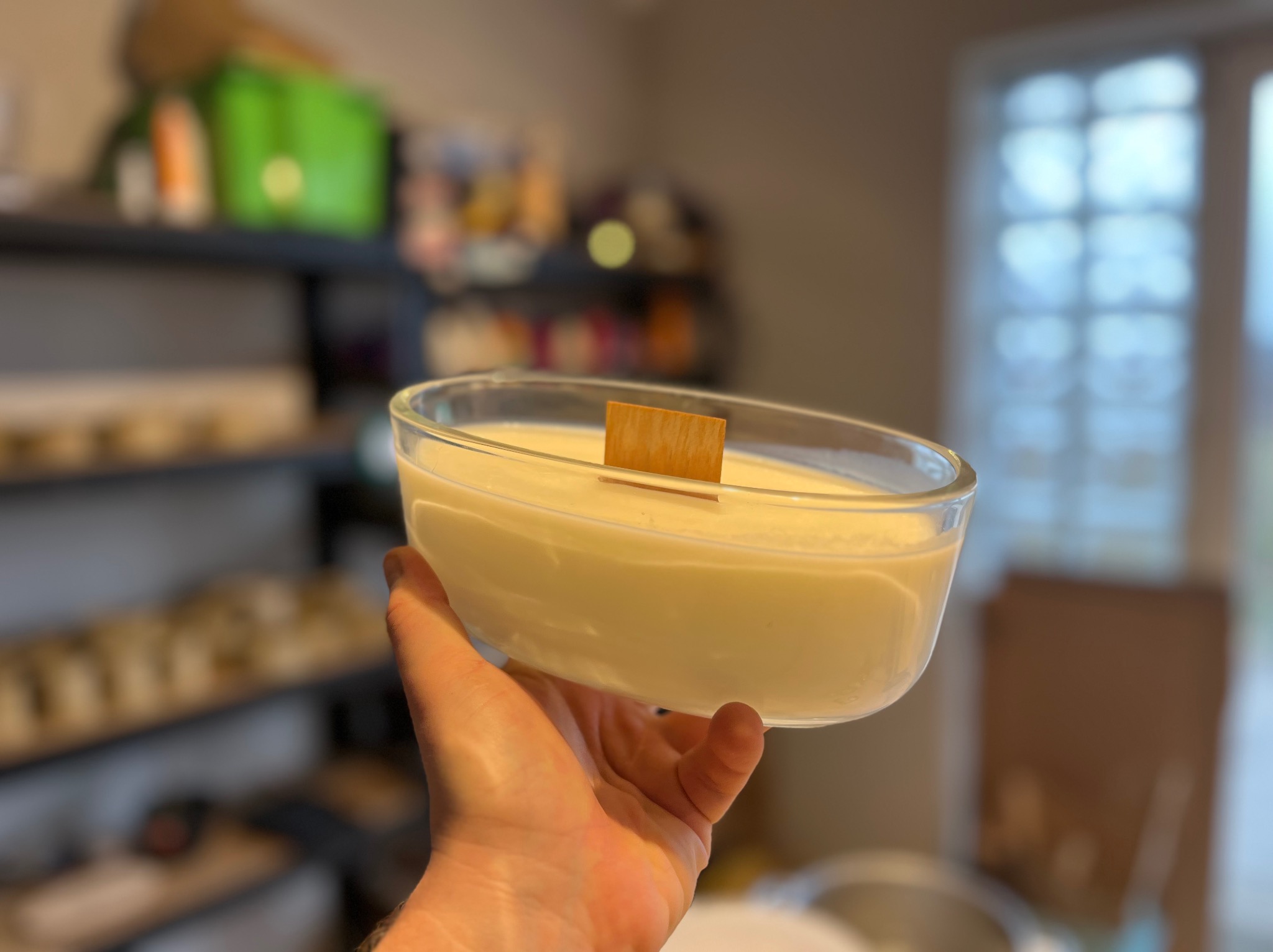 Ręka trzyma szklane naczynie wypełnione kremowym woskiem z drewnianym knotem, na rozmytym tle widać regał z różnymi przedmiotami i okno.