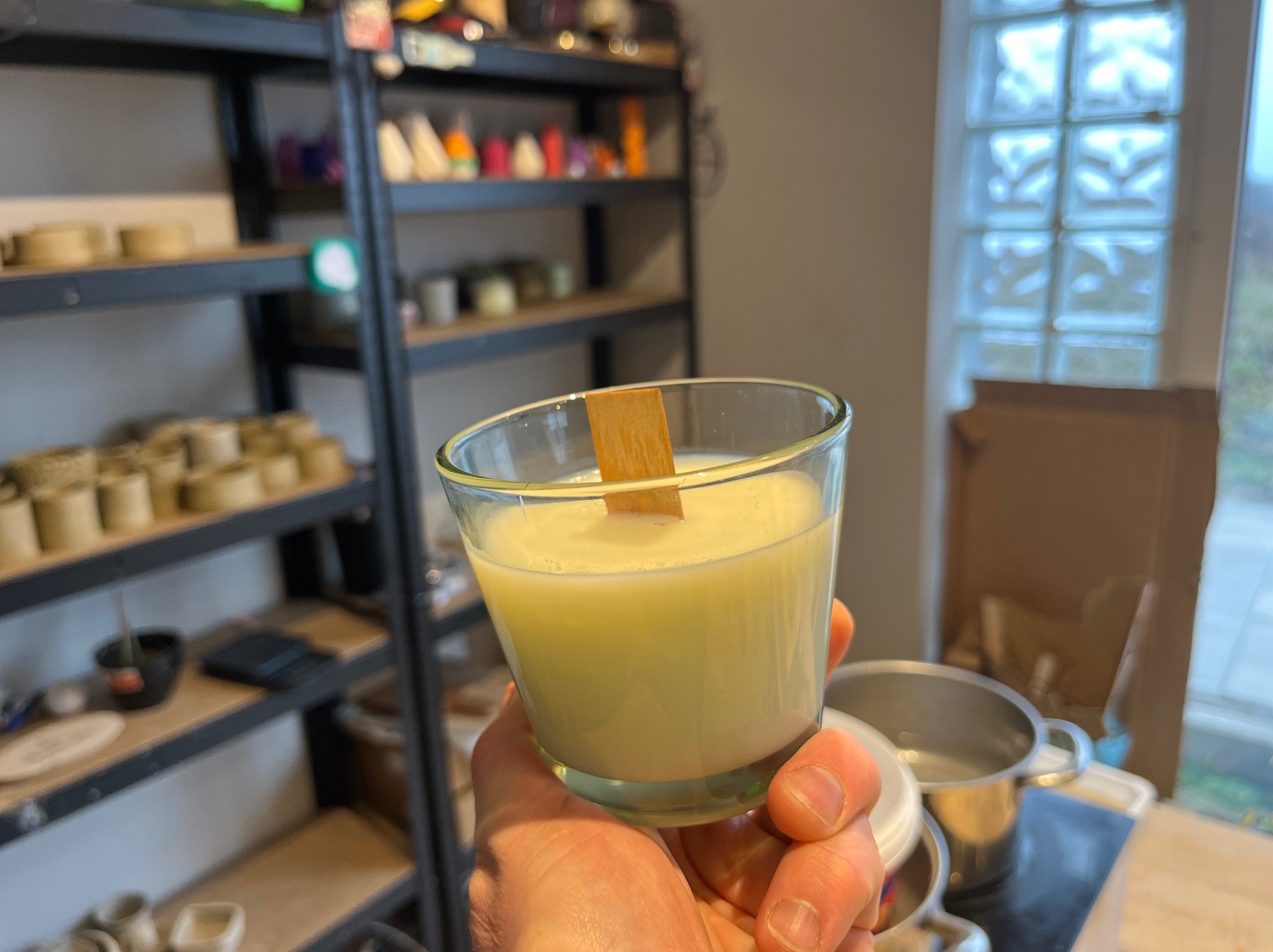 Ręka trzyma świecę sojową w szklanym naczyniu z drewnianym knotem, w tle warsztat z półkami pełnymi materiałów i narzędzi.