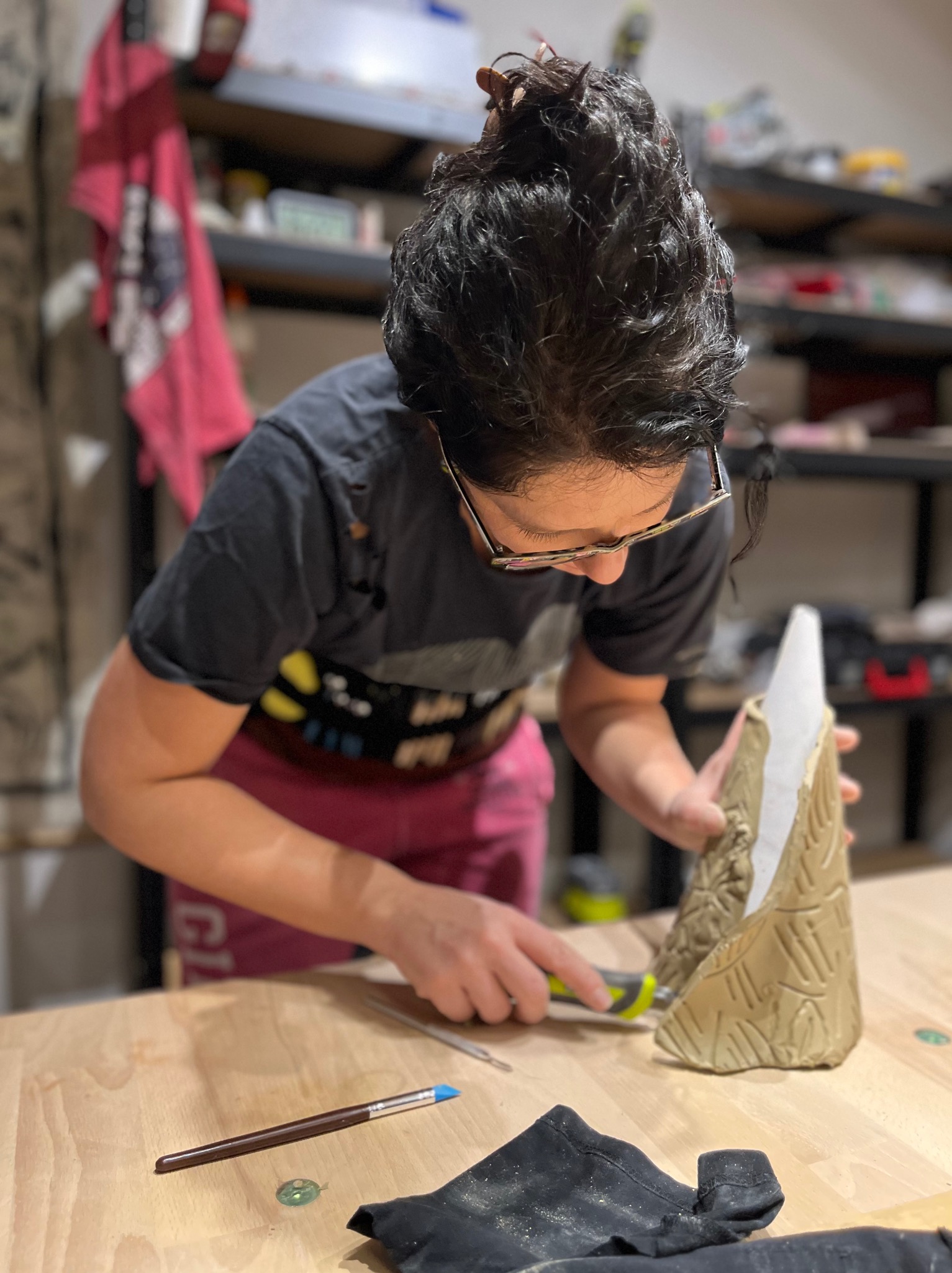 Artystka w Wilkasach rzeźbi w glinie stożkową formę z wzorami, używając nożyka rzeźbiarskiego, na drewnianym stole roboczym.